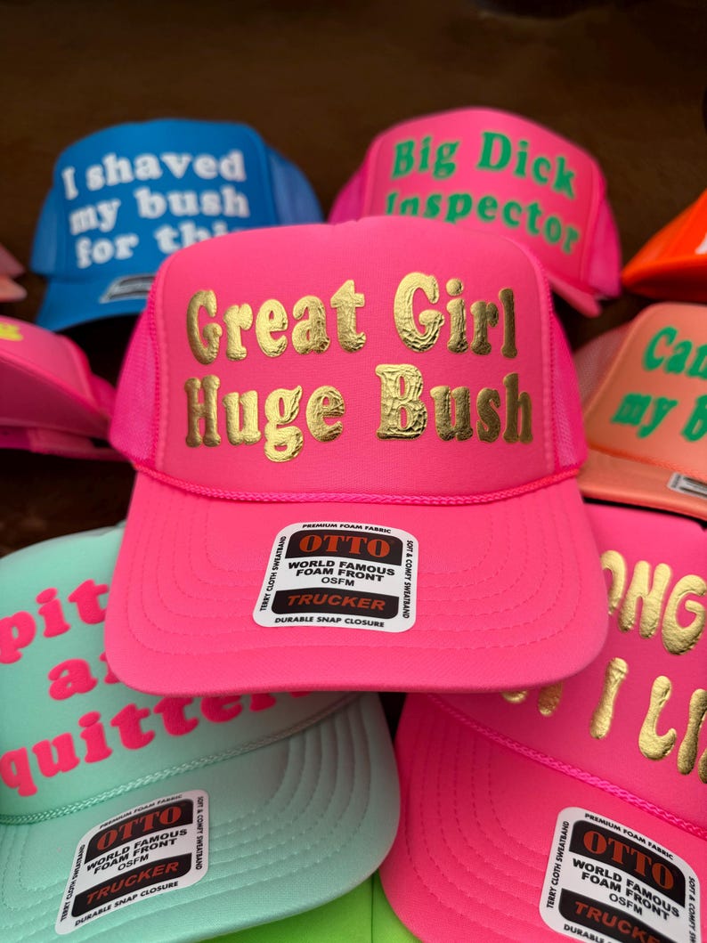 May include: A selection of trucker hats in vibrant colours like pink, blue, and green. The hats display text in gold and white, including phrases like "Great Girl Huge Bush" and "I shaved my bush for the". Each hat has a foam front and a snap closure.