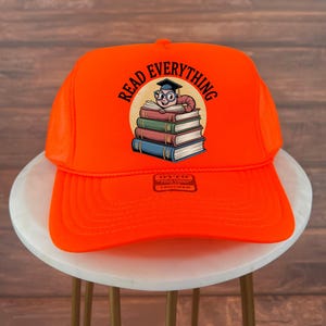 May include: Bright orange trucker hat with the words "READ EVERYTHING" in a circular design. The graphic features a cartoon worm wearing glasses and a graduation cap, sitting atop a stack of books.