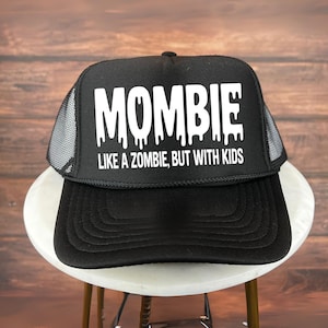 Könnte beinhalten: Schwarze Trucker-Mütze mit weißem Text, der "MOMBIE LIKE A ZOMBIE, BUT WITH KIDS" lautet. Die Mütze hat einen Mesh-Rücken und einen gebogenen Schirm. Die Mütze liegt auf einer weißen Oberfläche.