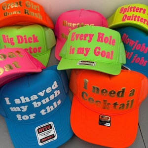 May include: A selection of trucker hats in vibrant colours such as neon green, pink, blue, and orange. Each hat displays striking text in contrasting colours, with phrases like "Every Hole is my Goal" and "I need a Cocktail."