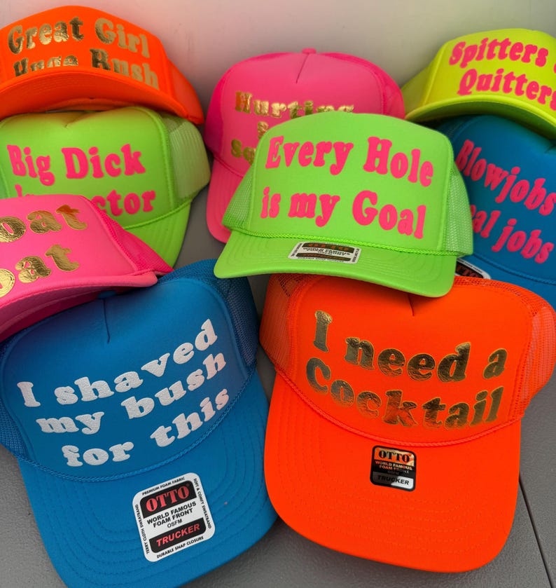 May include: A selection of trucker hats in vibrant colours such as neon green, pink, blue, and orange. Each hat displays striking text in contrasting colours, with phrases like "Every Hole is my Goal" and "I need a Cocktail."