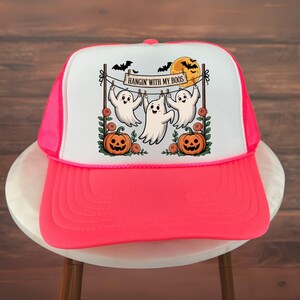 以下が含まれることがあります： ハロウィーンをテーマにした、ネオンピンクと白のトラッカーハット。 グラフィックには、「HANGIN' WITH MY BOOS」の文字が入った物干しロープにぶら下がっている3匹の漫画の幽霊、カボチャ、コウモリ、満月が描かれています。