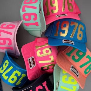 May include: A collection of colorful trucker hats with the number "1976" prominently displayed. The hats come in various colors, including pink, blue, and green, with contrasting text colors like gold and neon. The hats have a mesh back and a curved brim.