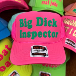 May include: A selection of trucker caps in vibrant neon shades, such as pink, green, and yellow. The caps display striking text in contrasting colours, with phrases like "Big Dick Inspector" and "Blowjobs are real jobs". Each cap has a foam front and a snap closure.