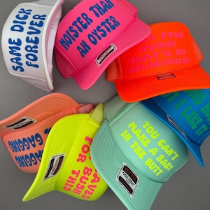 May include: A collection of trucker hats in various colors, including white, pink, orange, green, and blue. Each hat features bold text in contrasting colors. The phrases on the hats include "SAME DICK FOREVER" and "MOISTER THAN AN OYSTER."