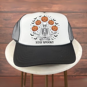 Puede incluir: Gorra de camionero en blanco y negro con diseño de Halloween. El gráfico presenta un esqueleto meditando con calabazas y murciélagos. El texto "STAY SPOOKY" está impreso debajo. Visera negra y parte trasera de malla.