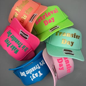 May include: A selection of brightly coloured trucker caps, each with a different slogan. The caps are in shades of pink, green, blue, and orange. Phrases include "Wake Pray It's Transfer Day," "Retrieval Day," and "Yay! It's Transfer Day."