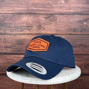 Op de afbeelding: Een marineblauwe baseballpet met een verstelbare band. De pet heeft een bruin leren patch met de tekst "YOUR TEXT/LOGO HERE" in oranje.