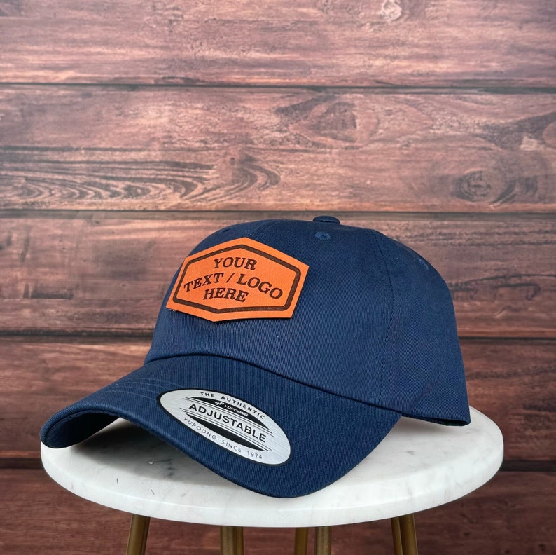 Op de afbeelding: Een marineblauwe baseballpet met een verstelbare band. De pet heeft een bruin leren patch met de tekst "YOUR TEXT/LOGO HERE" in oranje.