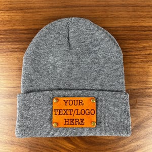 Puede incluir: Gorro de punto gris con un parche de cuero marrón que dice "YOUR TEXT/LOGO HERE".