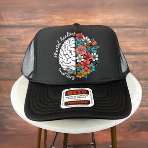 Peut inclure: Casquette de camionneur noire avec un dos en filet et une visière incurvée. L'avant présente un dessin d'un cerveau et de fleurs avec les mots "mental healing matters". La casquette a une fermeture à pression et un devant en mousse.