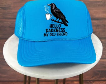 May include: A bright blue trucker hat with a black raven perched on a branch, holding a coffee cup. The hat features the text "HELLO DARKNESS MY OLD FRIEND" in black. The hat has a mesh back and a curved brim.