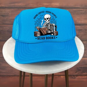 Op de afbeelding: Een felblauwe truckerpet met een witte voorkant. De pet heeft een afbeelding van een skelet dat een boek leest, met de tekst "DRINK COFFEE - AVOID PEOPLE" en "READ BOOKS".