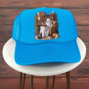 Op de afbeelding: Felblauw truckerpetje met een afbeelding van een lezende geest in een stoel, omringd door boeken, een kat, een pompoen en een schedel. De pet heeft een mesh achterkant en een gebogen klep.