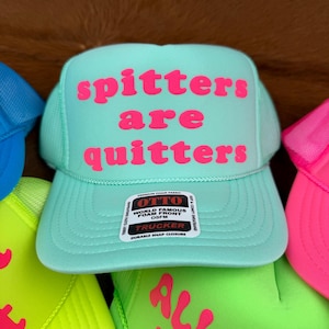 May include: A turquoise trucker hat with the phrase "spitters are quitters" in pink lettering. The hat features a mesh back and a snap closure. Other hats with text are visible.
