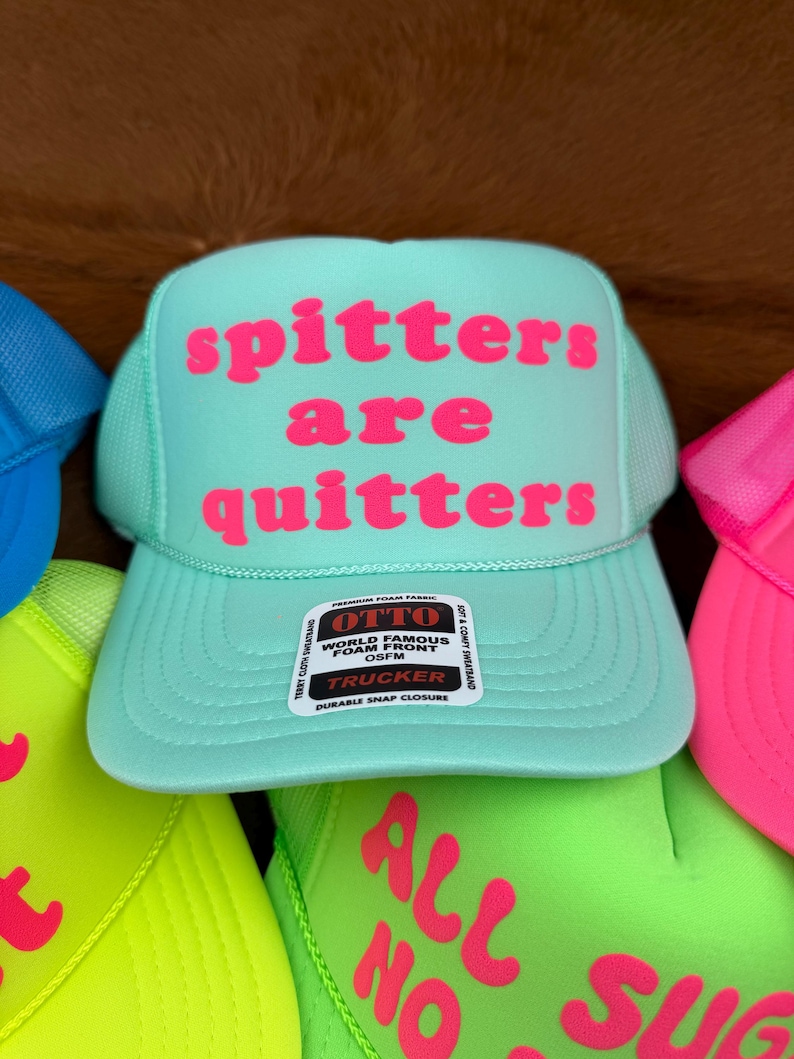 May include: A turquoise trucker hat with the phrase "spitters are quitters" in pink lettering. The hat features a mesh back and a snap closure. Other hats with text are visible.