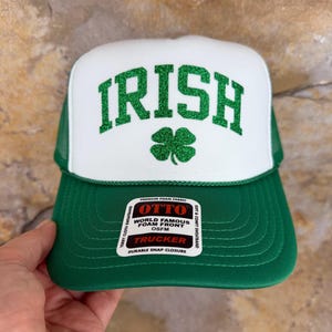 May include: Green and white trucker hat with the word "IRISH" in green glitter letters and a green shamrock. The hat has a green brim and a white front panel. The hat has a mesh back and a snap closure. The hat is labeled "OTTO".
