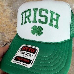 May include: Green and white trucker hat with the word "IRISH" in green glitter lettering and a green shamrock. The hat has a green brim and mesh back. The hat has a "TRUCKER" label.