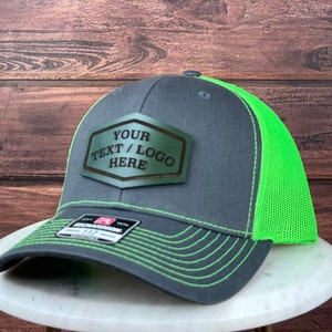 Könnte beinhalten: Eine graue und neongrüne Trucker-Kappe mit einem braunen Leder-Patch, auf dem "YOUR TEXT / LOGO HERE" steht.