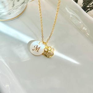 May include: A gold chain necklace with a white pearl pendant and a gold paw print charm with clear crystals. The pearl pendant has the letter "M" engraved on it.