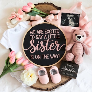 May include: A black and white round sign with the text "We are excited to say a little sister is on the way!" in pink lettering. The sign is surrounded by pink and white flowers, a pink teddy bear, a baby onesie, and a small wooden sign with the text "Baby Amelia Due Sept 2025".