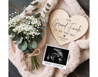 May include: A wooden heart-shaped sign with the text "OUR Grand Finale IS ON THE WAY. DECEMBER 2025" sits on a soft, beige blanket. A black and white sonogram photo with the text "BABY SIMONE" is next to the sign. A bouquet of white flowers and eucalyptus is tied with twine.