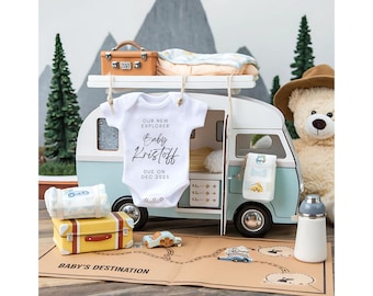 Può includere: Un body bianco con la scritta "Our New Explorer Baby Kristoff Due on Dec 2023" appeso a un camper blu e bianco in miniatura. Il camper è circondato da una mappa marrone, un orsacchiotto di peluche e una valigia gialla.