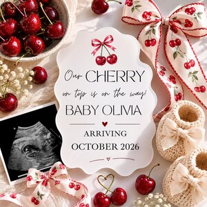 May include: A white scalloped sign with red cherry illustrations and text reading Our CHERRY on top is on the way! BABY OLIVIA ARRIVING OCTOBER 2026. Accompanied by a bowl of fresh red cherries, a sonogram, cream-colored baby booties, and cherry-print ribbon.