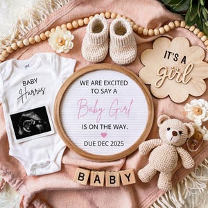 Może przedstawiać: Różowo-biały śpioszek dla dziecka z napisem "BABY Harris" i obrazem USG. Drewniany znak z napisem "WE ARE EXCITED TO SAY A Baby Girl IS ON THE WAY. DUE DEC 2025" oraz drewniany znak z napisem "IT'S A girl". Pluszowy miś szydełkowy i drewniane klocki układające się w słowo "BABY".