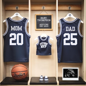 May include: Three basketball jerseys hanging on hooks in a locker room. The jerseys are navy blue with white lettering. The top jersey says "MOM 20", the middle jersey says "FUTURE MVP", and the bottom jersey says "DAD 25". A chalkboard sign above the jerseys says "BABY THOMPSON ARRIVING DEC 2023".