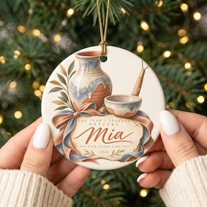May include: A round white ceramic ornament featuring a painted illustration of a pottery vase, a small bowl, and a tool. The text reads: THE YEAR I LEARNED POTTERY, Mia, YOUR STUDIO NAME, 2026. A gold cord is attached to the top for hanging purposes.