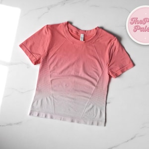 Puede incluir: Una camiseta de manga corta con degradado de coral a blanco. La camiseta presenta cuello redondo y un diseño entallado con paneles geométricos. Un logotipo circular con el texto "The Plain Palette" está en la esquina superior derecha.