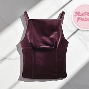 Puede incluir: Un top de terciopelo color ciruela con cuello cuadrado y tirantes finos. La prenda se exhibe sobre una superficie de mármol blanco con un logotipo circular rosa que dice "The Plain Palette".