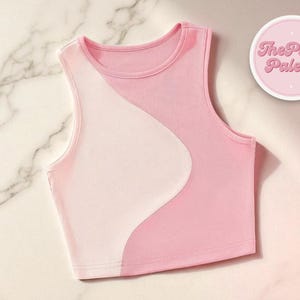 Könnte beinhalten: Ein ärmelloses, geripptes Crop-Top im Colorblock-Design. Das Top hat ein hellrosa und weißes Farbschema mit einem geschwungenen Design. Der Text "The Plain Palette" befindet sich in einem rosa Kreis oben rechts.