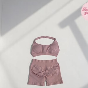 Puede incluir: Un conjunto de sujetador deportivo y shorts color malva a juego. El sujetador tiene un cuello halter y los shorts tienen un diseño fruncido en la espalda. El logotipo de The Plain Palette es visible en la esquina superior derecha.