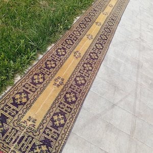 Op de afbeelding: Een lang, smal loperkleed met een centraal goudgeel paneel versierd met kleine, herhalende bloemmotieven. Het kleed is omlijst door brede, donkerpaarse randen met ingewikkelde geometrische patronen en gestileerde bloemvormen.