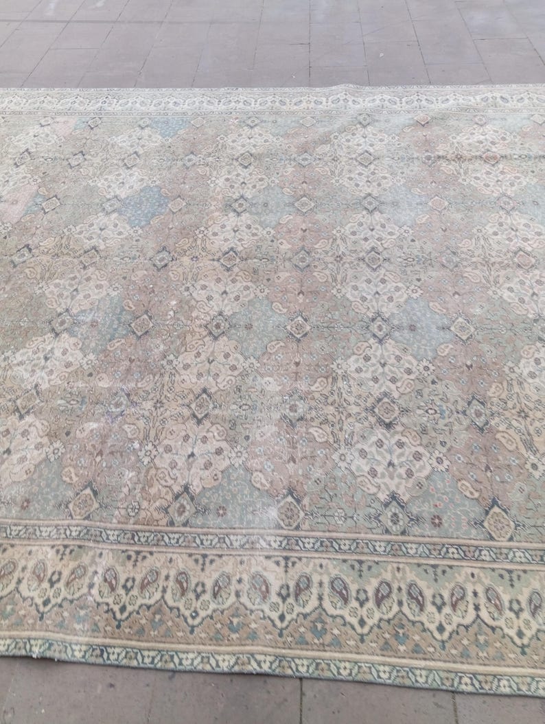 May include: A large, rectangular rug with an intricate, faded floral pattern in muted tones of beige, blue, and pale green. The rug features a detailed border with paisley-like motifs, creating a vintage aesthetic.