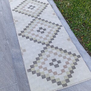 May include: A rectangular runner rug featuring an off-white base color with three repeating diamond-shaped geometric patterns. The diamonds are composed of small, pixelated squares in shades of muted olive green, charcoal gray, and light tan.