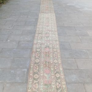 Puede incluir: Una alfombra larga y estrecha con un diseño tradicional. La alfombra presenta un patrón central de motivos florales rosas y verdes sobre un fondo beige, enmarcado por un borde de formas geométricas repetidas. La alfombra está colocada sobre una superficie de piedra gris.