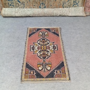 以下が含まれることがあります： ピンク、黒、黄色、クリーム色の幾何学模様が中央にある、小さな長方形のラグ。 ラグには繰り返し模様のボーダーがあり、灰色の表面に置かれています。 ロール状のラグが背景に見えます。