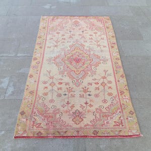 May include: Rectangular rug with a central medallion design in shades of pink, red, and beige. The rug features an intricate floral pattern and a border in a complementary color palette. The rug is placed on a gray stone surface.