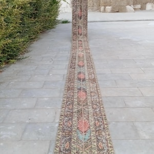 May include: A long, narrow, vintage runner rug with a detailed, repeating pattern. The rug features a central design of diamonds and floral motifs in shades of brown, peach, and blue. The rug is displayed outdoors on a stone surface.