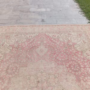 May include: A large rectangular area rug featuring a faded pink and beige colour palette. The rug displays an intricate, symmetrical floral pattern with a central medallion and detailed border motifs woven throughout the entire surface area.