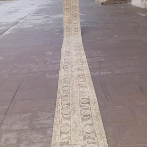 May include: A long, rectangular rug in cream and beige tones. The rug displays a detailed, repeating pattern with floral and geometric designs. The rug is shown outdoors on a concrete surface, highlighting its length and intricate design.