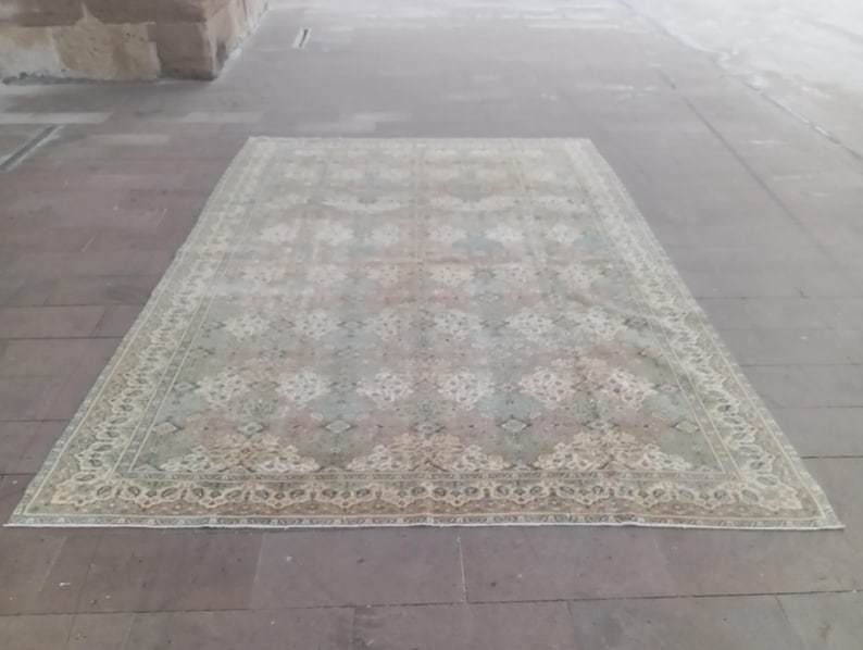 May include: A rectangular, faded-look rug with a detailed floral pattern in shades of beige, green, and blue. The rug has a decorative border and is placed on a stone surface.