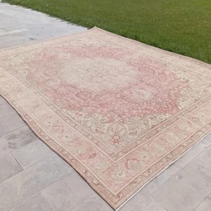 May include: A large rectangular area rug featuring a faded pink and cream colour palette with an intricate floral medallion pattern in the centre and a detailed border. The rug has a low pile texture and a symmetrical design across its surface.