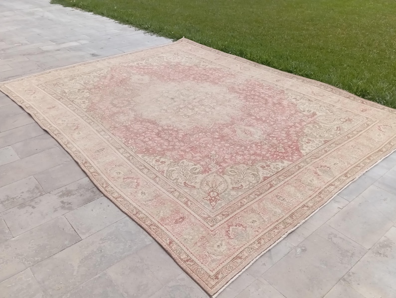 May include: A large rectangular area rug featuring a faded pink and cream colour palette with an intricate floral medallion pattern in the centre and a detailed border. The rug has a low pile texture and a symmetrical design across its surface.