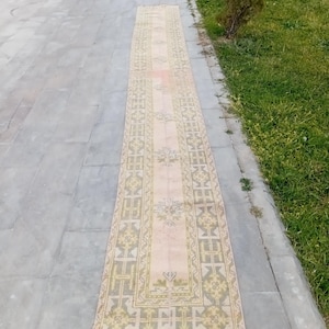 May include: A long, rectangular rug with a faded, vintage appearance. The rug features a central design in shades of pink and beige, framed by a border of green and yellow geometric patterns. The rug's edges are finished with a red trim.