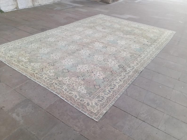 May include: A rectangular, vintage-style rug with a faded, distressed appearance. The rug features a central pattern in shades of pale green, pink, and blue, surrounded by a detailed border in cream and beige tones. The rug is placed on a wooden floor.