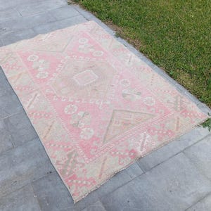 May include: A rectangular area rug featuring a faded pink and beige color palette with a central diamond medallion and intricate geometric floral motifs throughout the field, finished with a decorative border and short fringe along the edges.